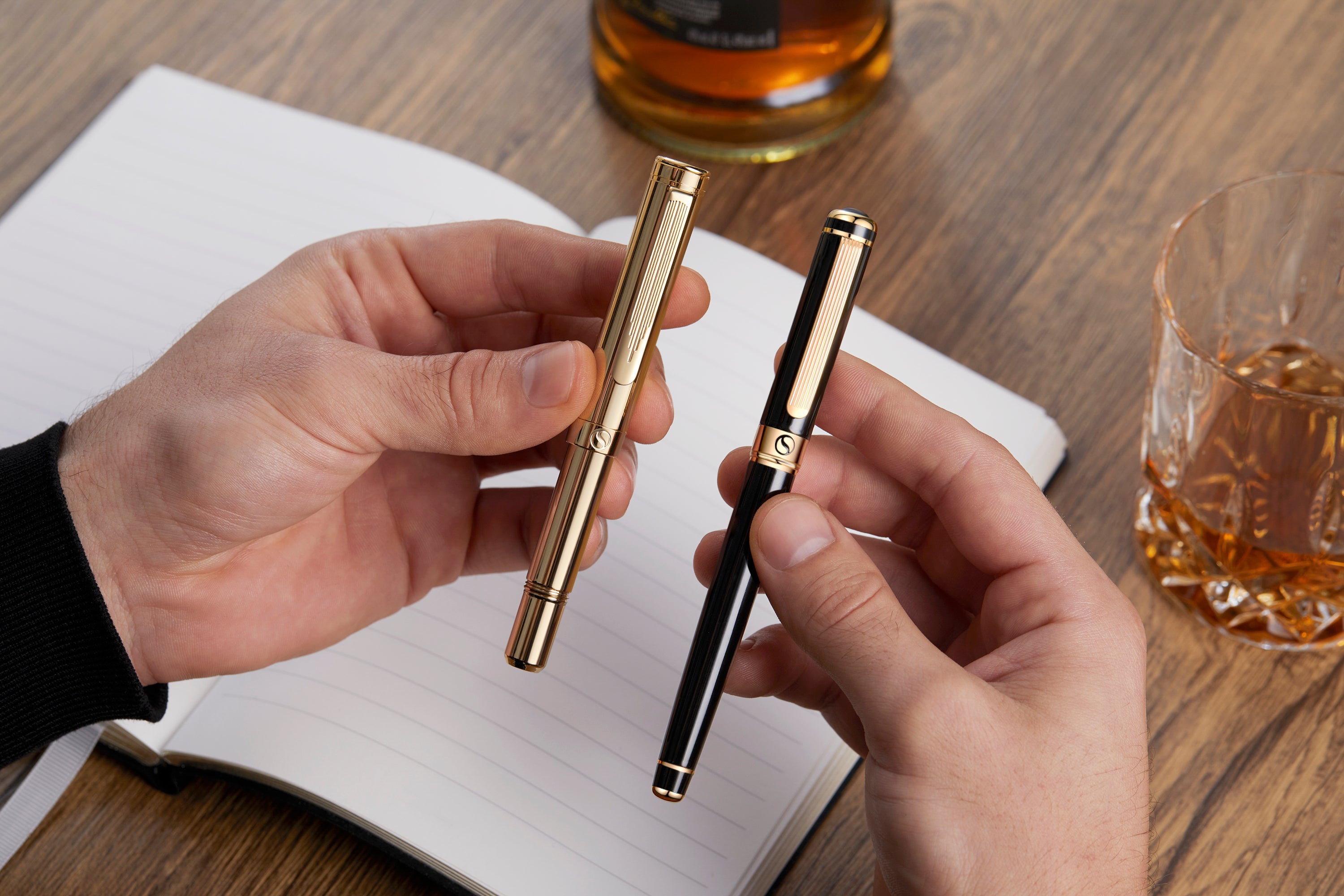 Two hands holding a gold and black pen on a wooden table with glasses of whiskey.