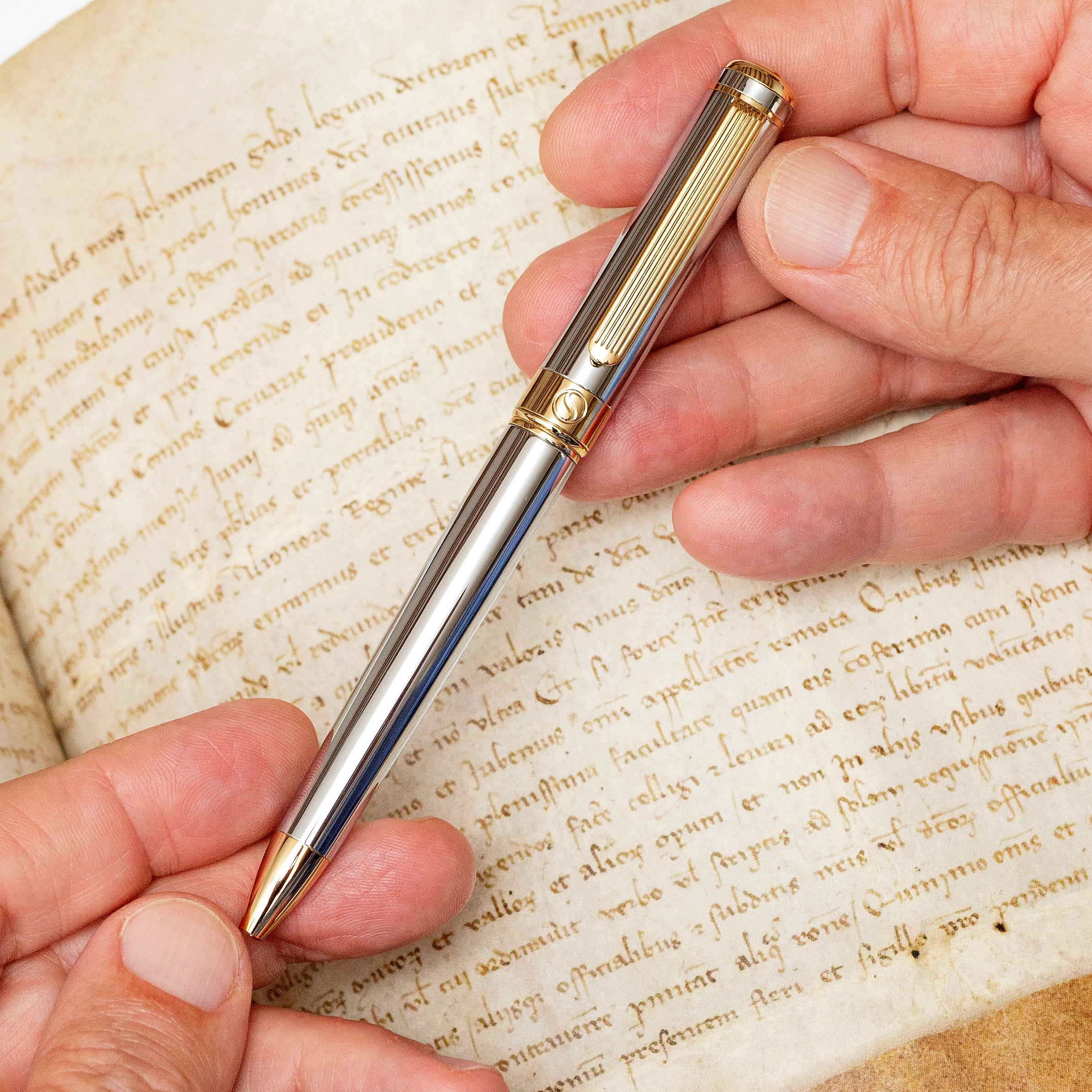 Hand holding a silver and gold pen over an old document with cursive writing.